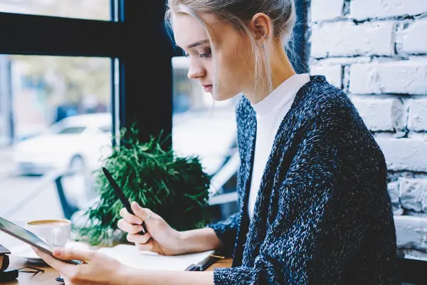 Modern güzel sarışın saç kadın yan görünüm masada kahve dükkanı pencere yakın tablet kullanarak ve Not defteri 'nde not alarak oturuyor. Hipster kız Blogger oluşturma Makale