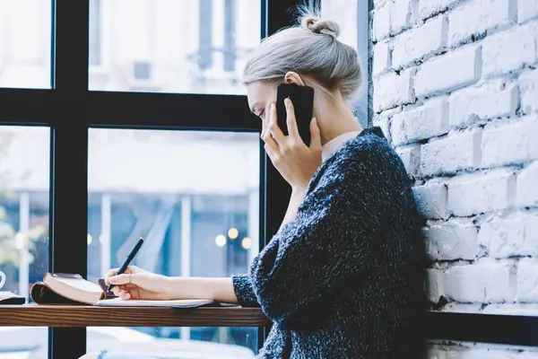 Modern Casual giyinik sarışın kadın kahve dükkanında masada oturan akıllı telefonla arama ve Not defteri 'nde serbest çalışırken notları alarak