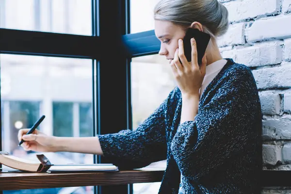 Kahve dükkanı iç masada otururken 20 yaşında kadın cep telefonu ile arama yan görünümü. Genç kadın Freelancer kullanarak teknoloji ve yazma notları Notepad