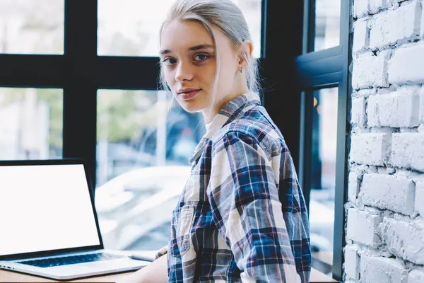 Gülümseyen genç Casual sarışın saç hipster kız kamera bakarak kafede pencere yakın masada ekran dizüstü bilgisayar ön Mock kadar oturuyor. Kahve dükkanı iç bilgisayar ile online öğrenme kadın öğrenci