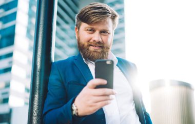 Resmi olarak başarılı bir adamın yarım boy portresi cep telefonu cihazını elinde tutuyor ve finans bölgesinde kameraya gülümsüyor. Muhabbet etmek için akıllı telefonlu pozitif sakallı bir patron.