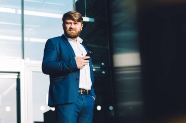 Elinde cep telefonu olan ciddi düşünceli bir erkek tüccarın tam boy portresi ofis girişimi yakınında duruyor ve kameraya bakıyor, akıllı telefon bekleyen bir şirket çalışanı.