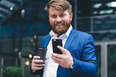 Neşeli erkek girişimcinin yarım boy portresi. Lezzetli kahve bardağıyla birlikte. İnternet sitesi, iş adamları kavramı ve finans haberlerini kontrol etmek için cep telefonu uygulaması kullanıyor.