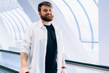 Yakışıklı hippi adamın tam boy portresi şık giyinmiş alışveriş merkezinde elektrikli merdivenlerde dikilip boş zamanlarında kameraya bakıyor, yürüyen merdiven ile ulaşım kavramına bakıyor.