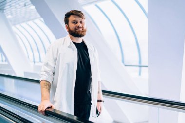 Yakışıklı hippi adamın tam boy portresi şık giyinmiş alışveriş merkezinde elektrikli merdivenlerde dikilip boş zamanlarında kameraya bakıyor, yürüyen merdiven ile ulaşım kavramına bakıyor.