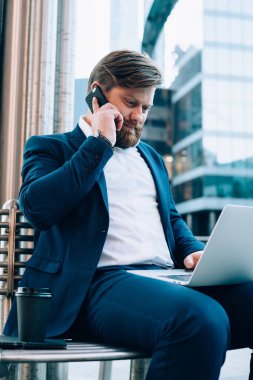 Zarif takım elbiseli erkek patron iş arkadaşıyla cep telefonu görüşmeleri sırasında dizüstü bilgisayara not alıyor. Akıllı telefon cihazıyla dengeyi kontrol etmek için servis operatörüyle konuşuyor.