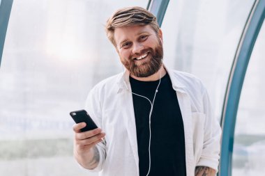 Elektronik kulaklıkla favori ses yayını dinlerken kameraya gülümseyen günlük kıyafetli mutlu sakallı melodinin yarım boy portresi, müzik eğlencesinden hoşlanan pozitif hipster adam.