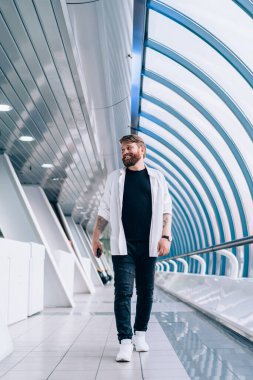 Pozitif sakallı bir adam, modaya uygun giyinmiş, halka açık bir yerde yürüyor ve gülümsüyor, neşeli bir beyaz erkek, varış yerine gidiyor, güzel görünümlü hippi çocuk.