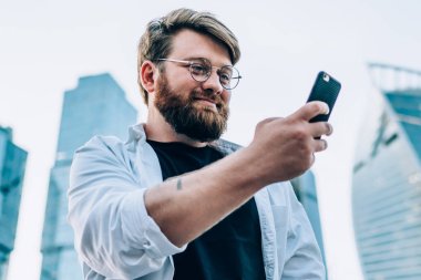 4G bağlantısını kullanarak cep telefonundan komik videolar izleyen, optik gözlüklü gülümseyen ve şehrin arka planında duran ağlardan iyi haberler okuyan genç adam.