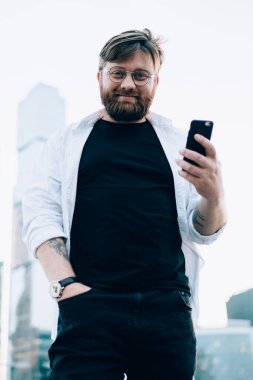 Metropolis 'in arka planında elinde cep telefonu cihazıyla duran mutlu erkek seyahat blogcusunun yarım boy portresi. Şehir caddesinde milenyum turisti kavramı olan kameraya bakıyor.