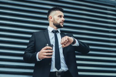 Sakallı müdür elinde bir fincan sıcak içecekle şehir caddesinde buluşmayı beklerken zamanı kontrol edip başka yere bakıyor.