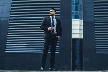 Çağdaş şehir caddesinde sıcak içeceğin tadını çıkarırken takım elbiseli, el ele tutuşmuş, gözlerini kaçıran dalgın sakallı bir erkek.