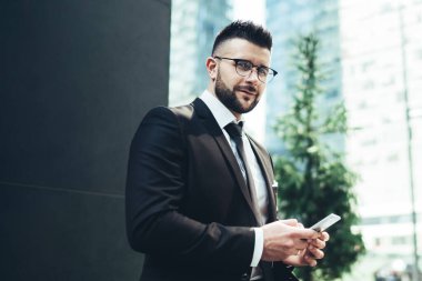 Şık siyah takım elbiseli mutlu yöneticinin bankta otururken kameraya gülümsemesi ve akıllı telefonu şehir merkezindeki bulanık gökdelenlere karşı kullanması.