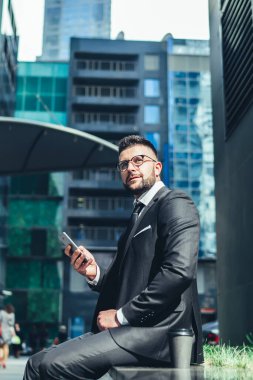 Kendinden emin, gözlüklü, şık elbiseli bir yönetici taştan bir bankta otururken ve modern binalara karşı kahve molası sırasında akıllı telefonla iletişim kurarken geleceğe yönelik planlar geliştiriyor.