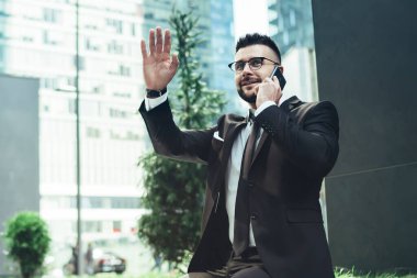 Gözlüklü ve resmi kıyafetli neşeli bir ofis çalışanının, taştan bir bankta oturmuş, şehir merkezinde gökdelenle telefonda konuşurken beşlik çakması.