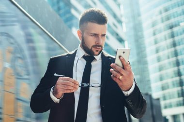 Moda tarzı iş kıyafetleri konusunda şaşırmış bir erkek yönetici cep telefonuyla sörf yaparken arka planda modern binalarla şehir merkezinde duruyor.