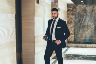 Siyah takım elbiseli ve kravatlı genç, zarif bir yönetici iş yerinde tek kullanımlık bir fincan kahveyle asansörü bekliyor.