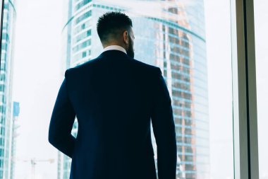 Yüzsüz, düşünceli, stil sahibi, takım elbiseli girişimcinin arka plandan görünüşü şehrin modern gökdelenlerinin ofisten bakışlarını kaçırması.