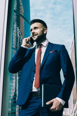 Mavi takım elbiseli, güneşli ofis odasında telefonla konuşan gökdelenlerin arkasından başka tarafa bakan olumlu içerikli adamdan.