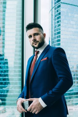 Dalgın, kendine güvenen, mavi takım elbiseli, akıllı telefonlu, çalışma alanındaki büyük cam ofis penceresine yaslanmış kameraya bakan modacı.