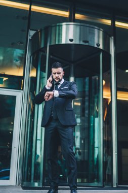 Takım elbiseli ciddi sakallı iş adamının düşük açılı görüntüsü akıllı telefondan konuşuyor ve şehir dışındaki modern binanın dışında buluşmadan önceki zamanı kontrol ediyor.