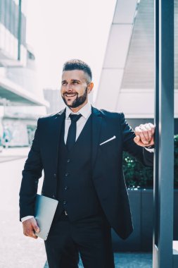 Elinde dizüstü bilgisayarı olan neşeli erkek yönetici şehir merkezinde başarılı bir iş adamı, şehir merkezinde başarılı bir iş adamı beklerken gülüyor ve eğleniyor.