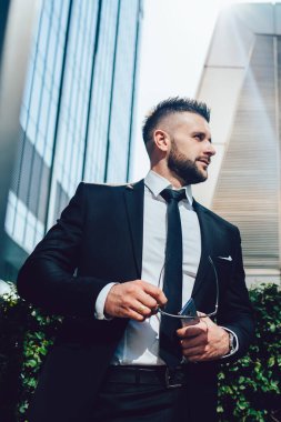 Düşük açılı, takım elbiseli, gözlüklü, akıllı telefonlu ve çalılıkların yanında dikilirken yüzünü çeviren başarılı sakallı erkek.