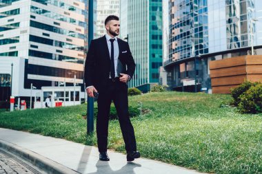 İş alanındaki modern şehir kulelerine karşı caddede yürüyen dosyasıyla kurumsal takım elbiseli içerikli profesyonel işçi başka yöne bakıyor.