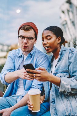 Akıllı telefondan uygulama gösteren Afrikalı Amerikalı genç bir kadın günlük kot giyen hippinin dışarıda kahveyle oturduğunu merak etti. Cep telefonundan video izleyen çok kültürlü arkadaşlar.