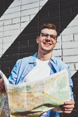 Gözlüklü neşeli beyaz adam elinde seyahat haritası tutarken ve duvarın karşısında duvarla dikilirken yüzünü çeviriyor. Günlük kıyafetleriyle gülen mutlu hippi turist.