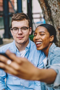 Neşeli afro-amerikan genç kadın akıllı telefonun önünde beyaz tenli hippi adamla birlikte poz vererek internet sitesinde otururken fotoğraf çekiyor. Çeşitli arkadaşlar selfie çekiyor.