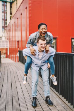 Mutlu Afrikalı Amerikalı hippi erkek gülüyor erkek arkadaşının omzuna oturuyor dışarıda eğleniyor, çok ırkçı çift birlikte vakit geçiriyor erkek kızı sırtında gezdiriyor