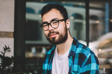 Sıradan kıyafetler ve gözlüklerle takılan sakallı etnik bir adam sokak restoranında masanın yanında otururken kameraya bakıyor.