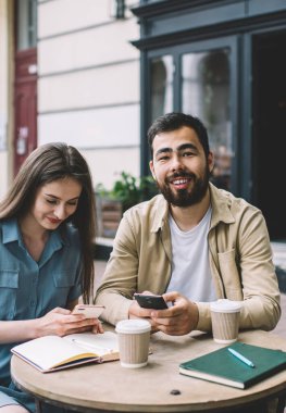 Etnik sakallı adam ve genç kadın, ara verildiğinde şehir sokaklarında restoranda otururken gülümseyen ve akıllı telefonlara bakıyorlar.