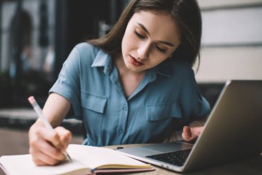 Çok çalışan genç esmer, gündelik kıyafetlerle dizüstü bilgisayarla çalışıyor ve açık bir kafede otururken notlar yazıyor.