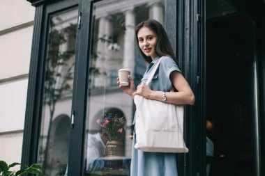 Modern, yoğun bir bayanın gündelik kıyafetlerini omuzlarında modaya uygun beyaz bir bez torbayla tek kullanımlık kahve bardağını tutarken ve kahve dükkanından çıkarken kameraya bakarken...