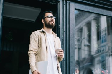 Dizüstü bilgisayarı ve tek kullanımlık kahveyi tutan ve yaz gününün tadını çıkaran serbest çalışan genç bir erkek kahvehanenin kapısında beklerken yaptığı işten memnun.
