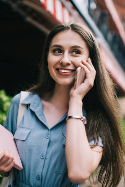 Not defteriyle mutlu genç bir bayan. Gülümsüyor ve uzağa bakıyor. Akıllı telefonla konuşuyor ve şehrin bulanık arka planında yürüyor.