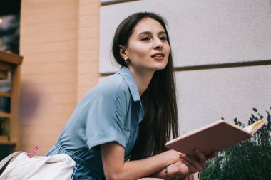 Mavi elbiseli rüya gibi genç bir öğrencinin şehir binasının önünde oturmuş not defteriyle notlar okurken gözlerini kaçırması.