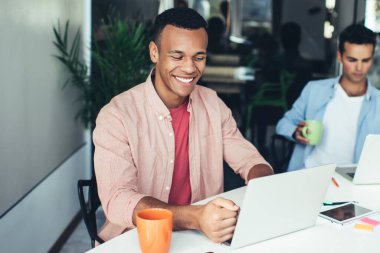 Mutlu Afrikalı Amerikalı erkek yazılım geliştirici modern laptop cihazıyla ofis masasında oturuyor ve komik video, teknoloji ve iletişim konsepti ile e-posta yoluyla gülümsüyor.
