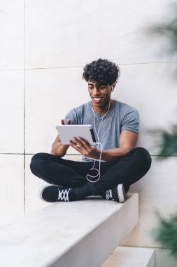 Elektronik kulaklık ve dokunmatik ped uygulaması ile eğlenceli müzik dinlemek için şehir merkezinde boş vakitlerin tadını çıkaran pozitif hipster adam, tabletle meloman arayan neşeli Latin adam web sitesi