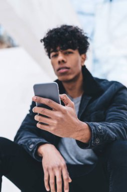 Bin yıllık Latin adamın portresi cep telefonu cihazını elinde tutuyor ve iletişim için halka açık internete bağlanan kameraya bakıyor, blog yazmak için dijital cep telefonuna seçici odaklanıyor