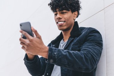 Cep telefonu cihazlı genç, olumlu hipster arkadaşı ile görüntülü iletişim kuruyor, mutlu milenyum erkek blogcusu akıllı telefon içeriğini okuyarak kentsel arka planda duruyor.