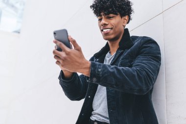 Cep telefonu cihazlı genç, olumlu hipster arkadaşı ile görüntülü iletişim kuruyor, mutlu milenyum erkek blogcusu akıllı telefon içeriğini okuyarak kentsel arka planda duruyor.