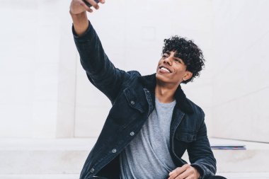 Hücresel aygıtlarla neşeli Latin bir adam akıllı telefon kamerasıyla selfie çekiyordu. Şehir merkezinde dinleniyordu. Sosyal ağlarda blog için video çeken mutlu genç bir erkek modern cep telefonu kullanıyordu.