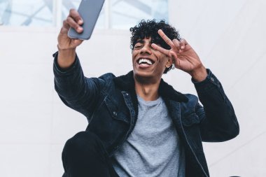 Neşeli hippi, modern akıllı telefonun ön kamerasında özçekim yapıyor. Bloglar için dışarıda zaman geçiriyor. Dijital cep telefonu aygıtı aracılığıyla Ziyaretçi işaretleri gösteren mutlu bir erkek.