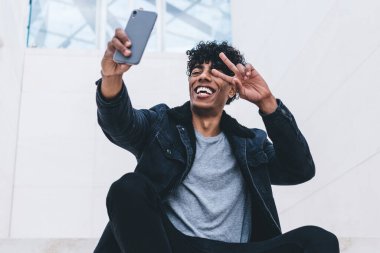 Neşeli hippi, modern akıllı telefonun ön kamerasında özçekim yapıyor. Bloglar için dışarıda zaman geçiriyor. Dijital cep telefonu aygıtı aracılığıyla Ziyaretçi işaretleri gösteren mutlu bir erkek.