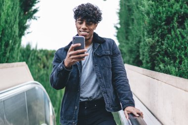 Şehre yürüyen merdivenden inen mutlu hipster adamı takipçilerle çevrimiçi iletişim kurmak için modern cep telefonu aygıtını kullanıyor. Sosyal sitede mesajlaşırken yakışıklı bir erkek blogger ağı kuruyor.