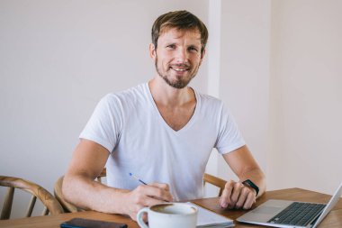 Gündelik beyaz tişörtlü başarılı erkek grafik tasarımcısının portresi kameraya gülümsüyor. Yeni web projesiyle uzaktan çalışmak için, halka açık bir kafede otururken, dizüstü bilgisayarı için.