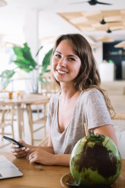 Modern cep telefonu ve dizüstü bilgisayarlı pozitif Kafkas dijital göçebelerinin portresi Bali kafeteryasında hindistan cevizi içecekleriyle iş için zaman harcıyor, kameraya gülümseyen mutlu serbest çalışan